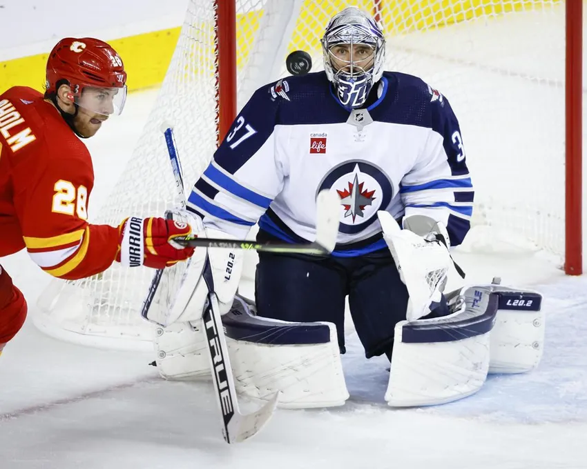 Le gardien des Jets de Winnipeg Connor Hellebuyck, à droite, suit la rondelle alors que l'attaquant des Flames de Calgary Elias Lindholm tente d'y mettre son bâton pendant la troisième période d'une action de hockey présaison de la LNH à Calgary, le vendredi 7 octobre 2022.